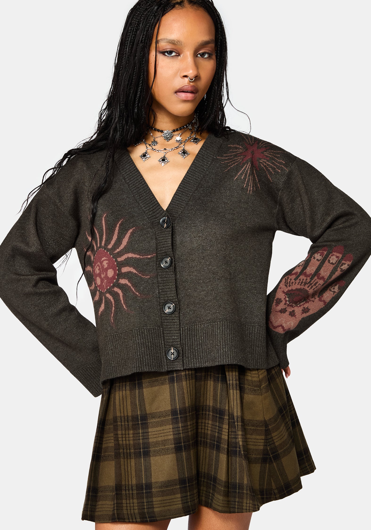 A woman modeling a dark brown button-up cardigan with red sun and handprint graphics, paired with a brown plaid pleated skirt, against a light gray background.