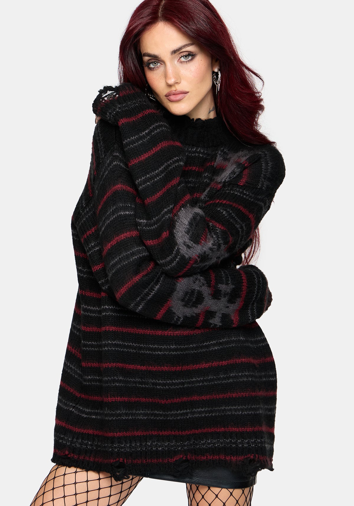 A woman with red hair wearing a black knit sweater dress with red and gray horizontal stripes, paired with black fishnet tights, against a light gray background.