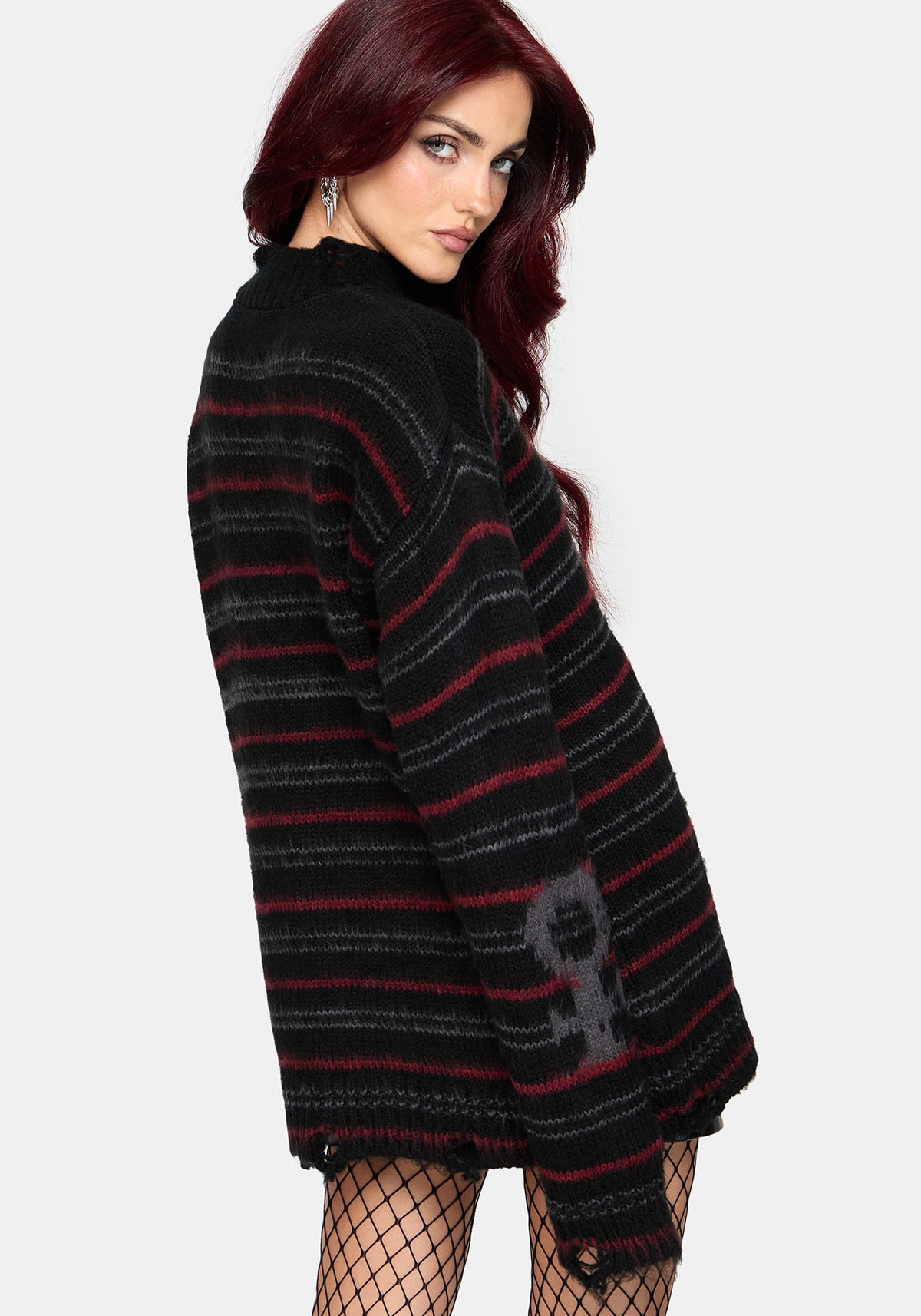 A woman with dark red hair models an oversized black knit sweater with thin red and gray horizontal stripes and a gray skull graphic on the sleeve, paired with fishnet stockings against a light gray studio background.