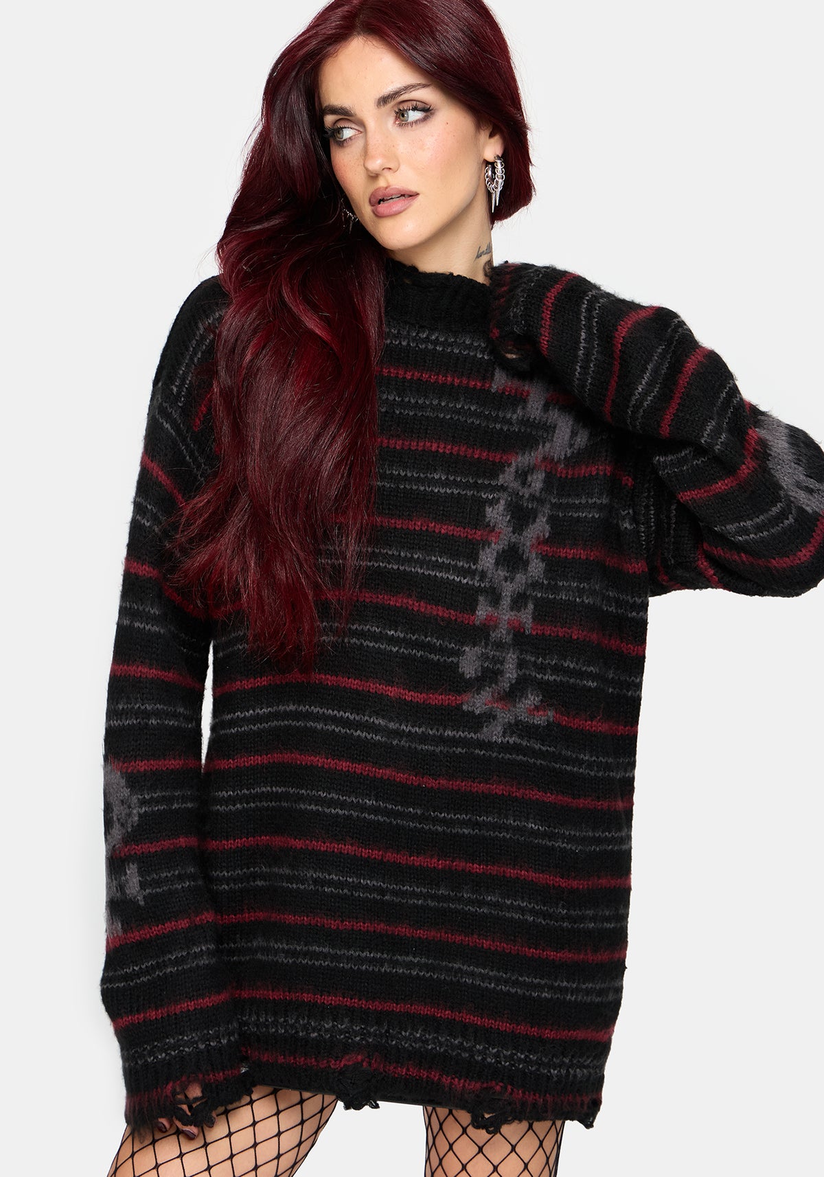 A woman with long dark red hair wearing a black and red striped knit sweater with distressed hem details and fishnet tights, posed against a light gray studio background.