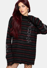 A woman with long dark red hair wearing a black and red striped knit sweater with distressed hem details and fishnet tights, posed against a light gray studio background.