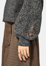 Close-up of a person wearing a dark gray marled knit sweater with a brown embroidered floral detail on the sleeve, paired with a brown and beige plaid skirt, with rings on the hand.