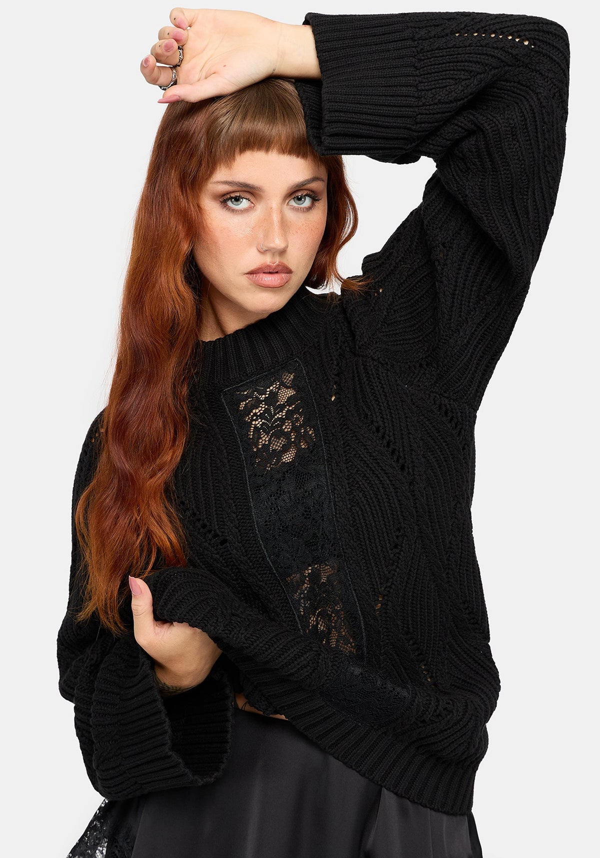 A woman modeling a black ribbed knit sweater with black lace panel details on the front, paired with a black skirt, against a light gray studio background.