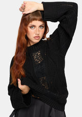 A woman modeling a black ribbed knit sweater with black lace panel details on the front, paired with a black skirt, against a light gray studio background.