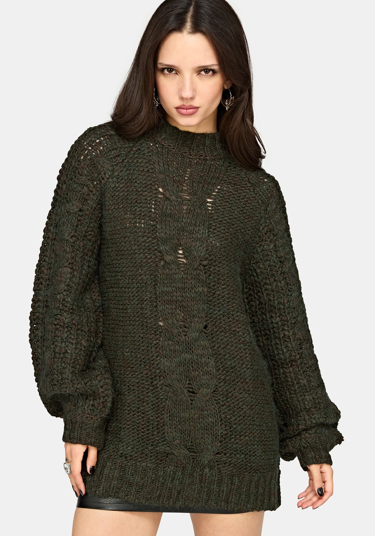 A woman modeling a dark olive green chunky knit sweater with distressed details, photographed against a light gray studio background.