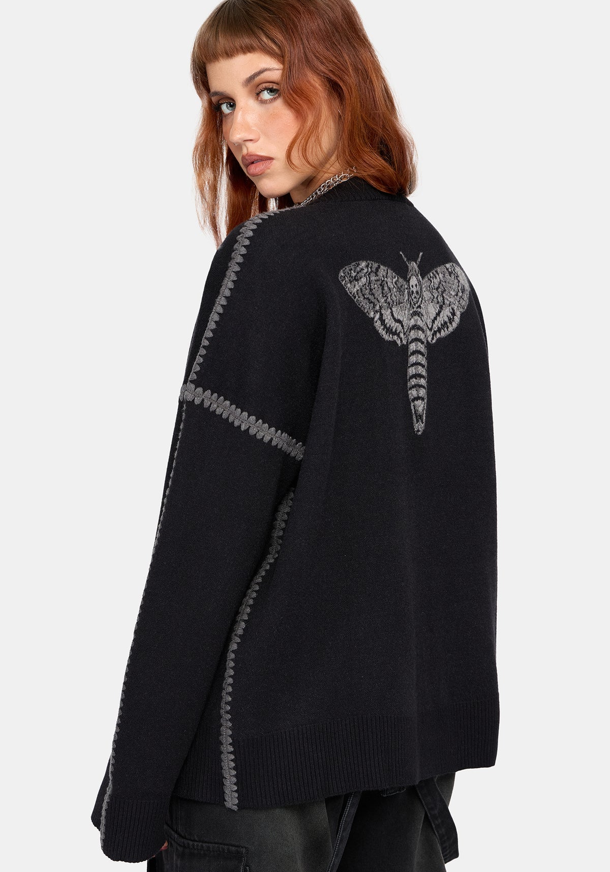 A woman wearing a black knit cardigan with gray contrast stitching and a large gray moth graphic on the back, photographed against a light gray studio background.