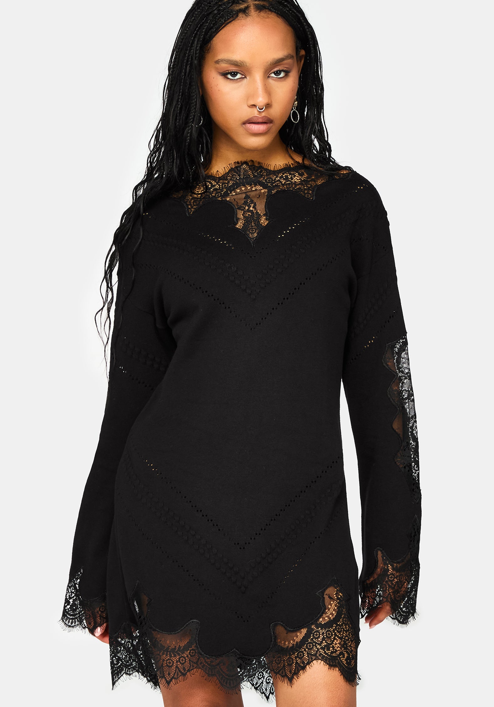 A woman modeling a black long-sleeve mini dress with intricate lace detailing at the neckline, cuffs, and hem, set against a plain light gray background.