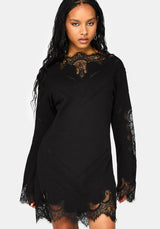 A woman modeling a black long-sleeve mini dress with intricate lace detailing at the neckline, cuffs, and hem, set against a plain light gray background.