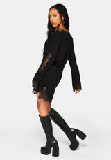 A woman modeling a black long-sleeve mini dress with sheer lace panels on the sleeves and hem, paired with black knee-high platform boots against a light gray studio background.