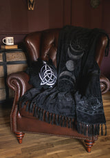 A black throw blanket and matching pillow set styled on a brown leather armchair in a cozy room. The blanket features white celestial designs including a sun, crescent moon, and full moon, along with fringe detailing.