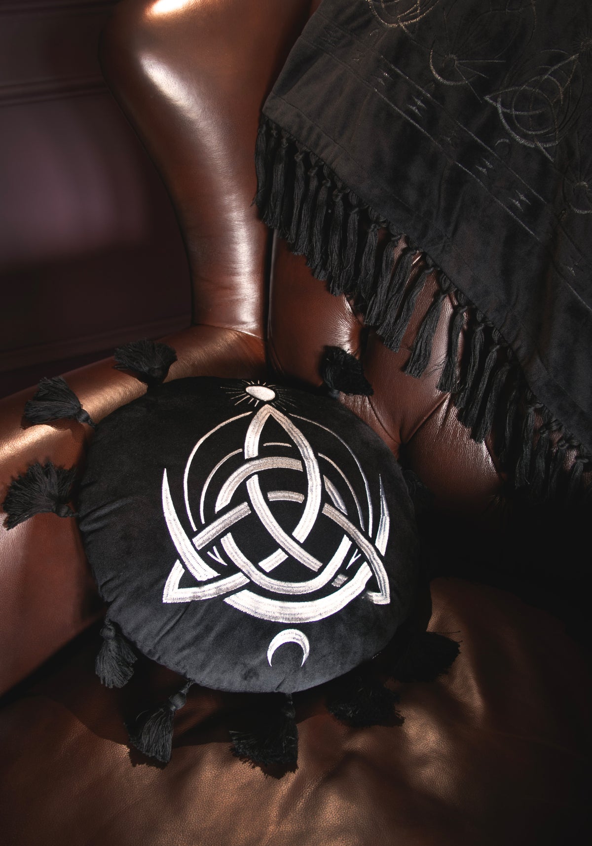 A circular black cushion with a white Celtic knot triquetra design, a small sun motif at the top, and tassels extending from the outer edge, shown on a light gray background.