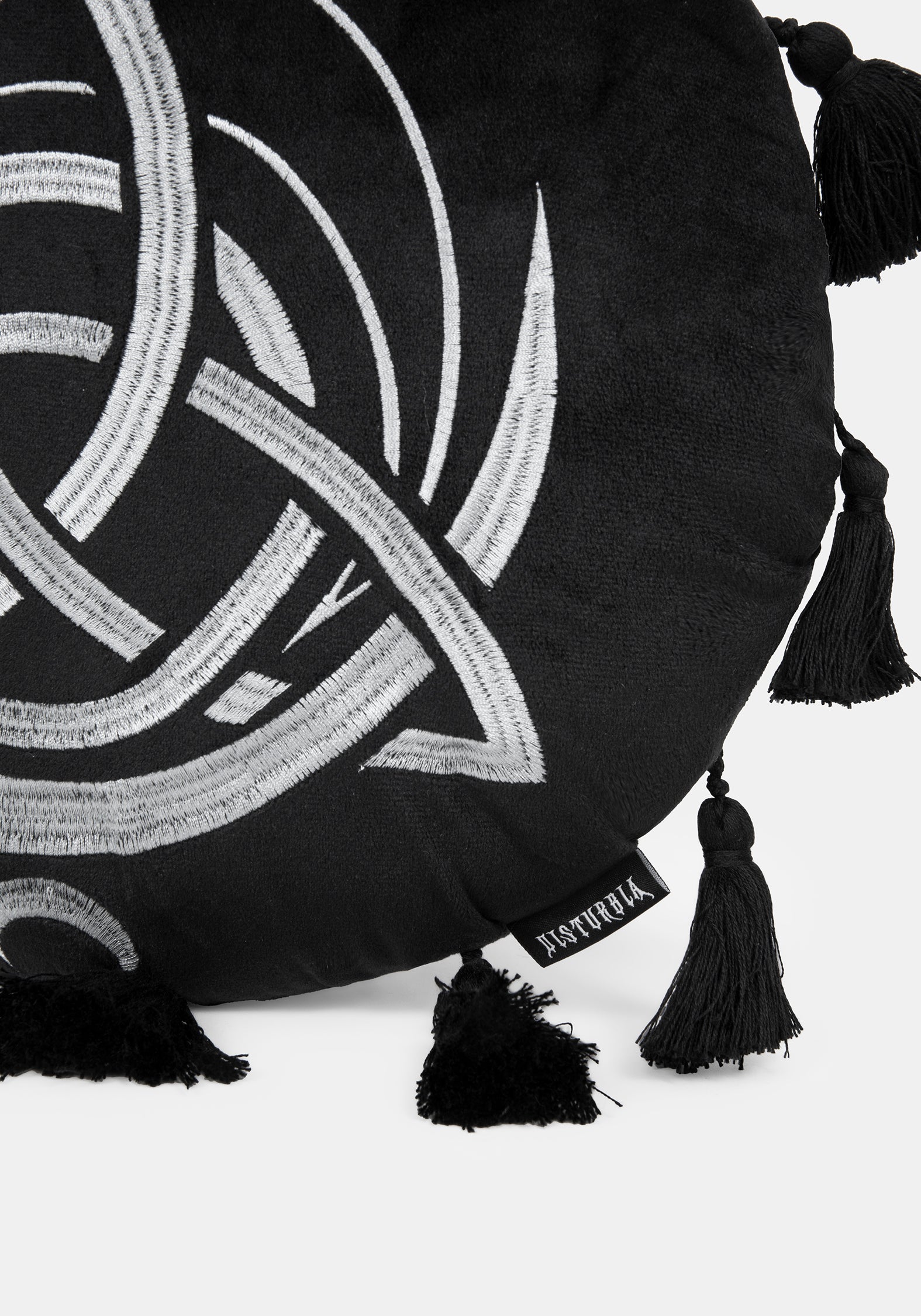 A circular black cushion with a white Celtic knot triquetra design, a small sun motif at the top, and tassels extending from the outer edge, shown on a light gray background.