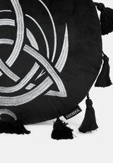 A circular black cushion with a white Celtic knot triquetra design, a small sun motif at the top, and tassels extending from the outer edge, shown on a light gray background.