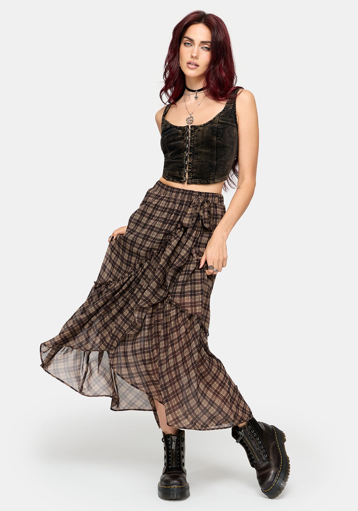 A model with dark red hair poses in a black velvet crop top and a brown plaid maxi skirt with black combat boots, set against a plain light gray background.