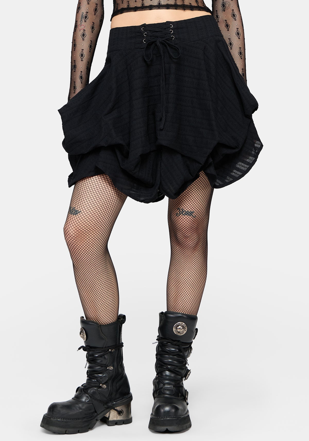 A model from the waist down wearing a black layered ruffled mini skirt with a lace-up front, paired with black fishnet tights and black lace-up combat boots, against a light gray studio background.