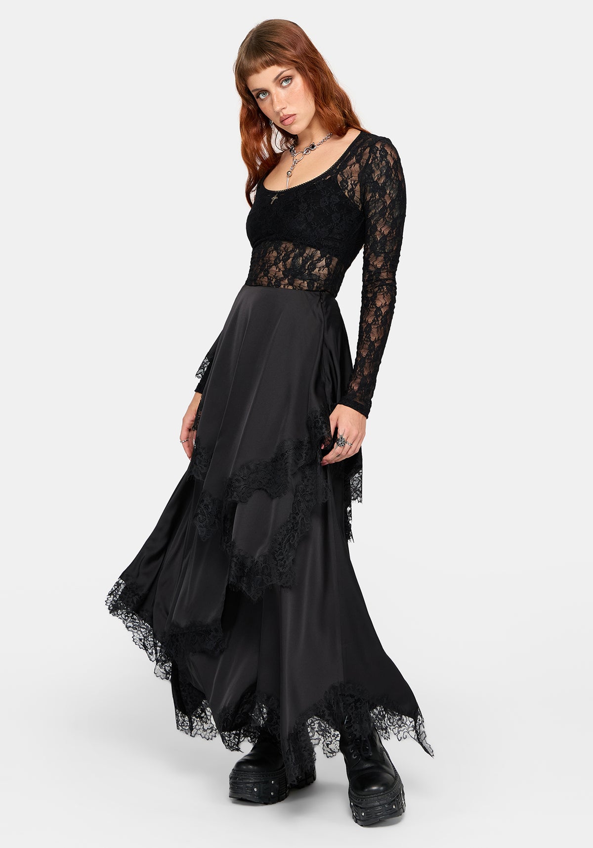 A model wearing a long black lace and mesh maxi dress with a high-low layered skirt and black platform boots, photographed against a light gray studio background.