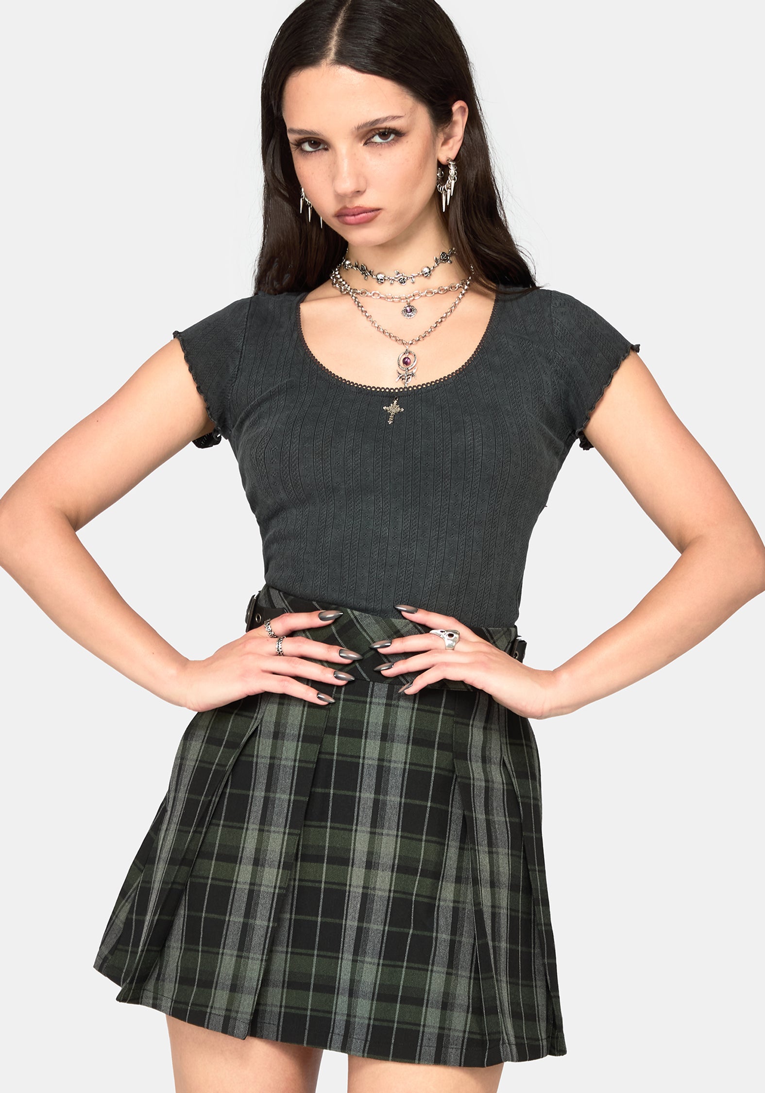 A woman posing in a dark gray ribbed short-sleeve top and a green and black plaid pleated mini skirt, accessorized with layered silver necklaces and hoop earrings, against a light gray background.