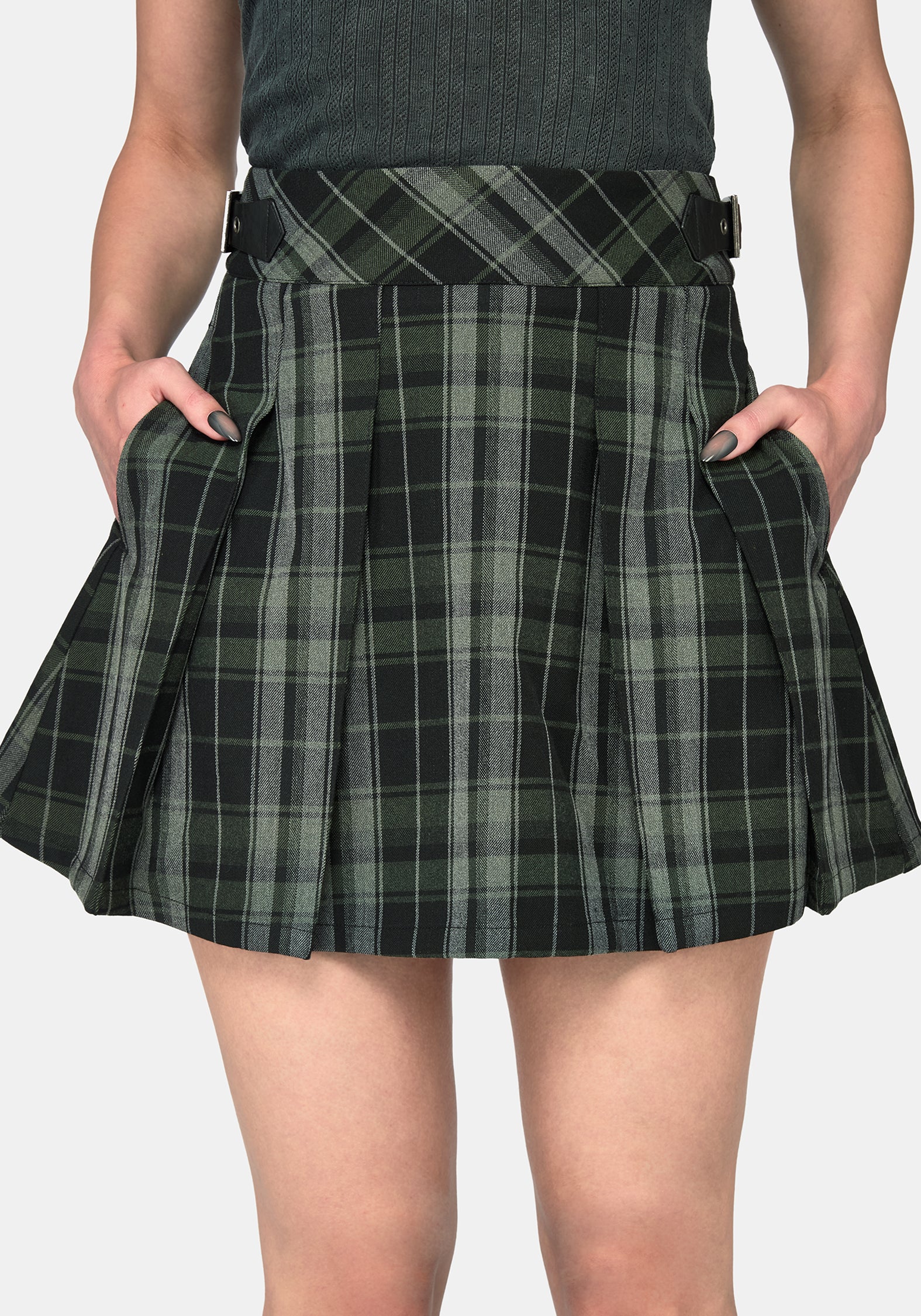 A close-up of a model wearing a pleated mini skirt in a green and black plaid pattern, with hands in the pockets, against a light gray background.