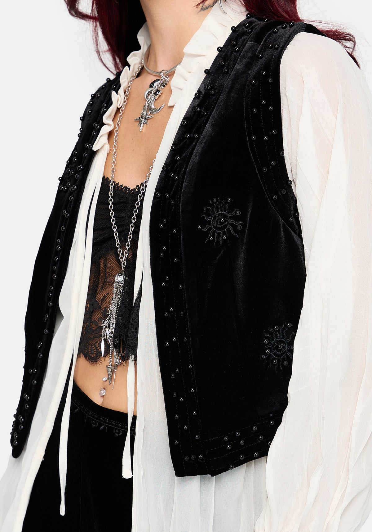 A close-up of a person wearing a black velvet vest with silver studs and embroidered sun motifs over a white ruffled shirt and black lace top, accessorized with layered silver necklaces.