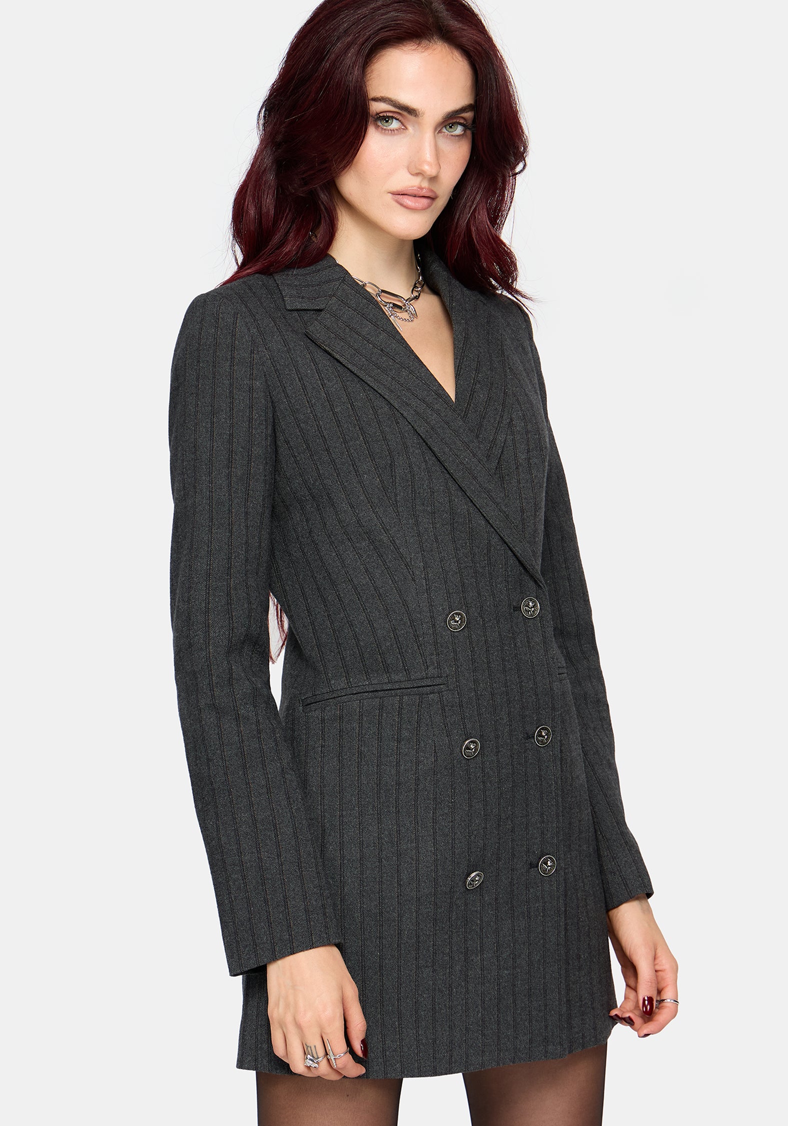 A woman wearing a dark charcoal gray double-breasted pinstripe blazer dress, shown from the waist up against a light gray studio background.