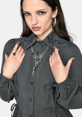 A close-up of a woman wearing a dark gray pinstriped shirt with black floral embroidery on the collar, styled with a silver heart-shaped pendant necklace and silver earrings.