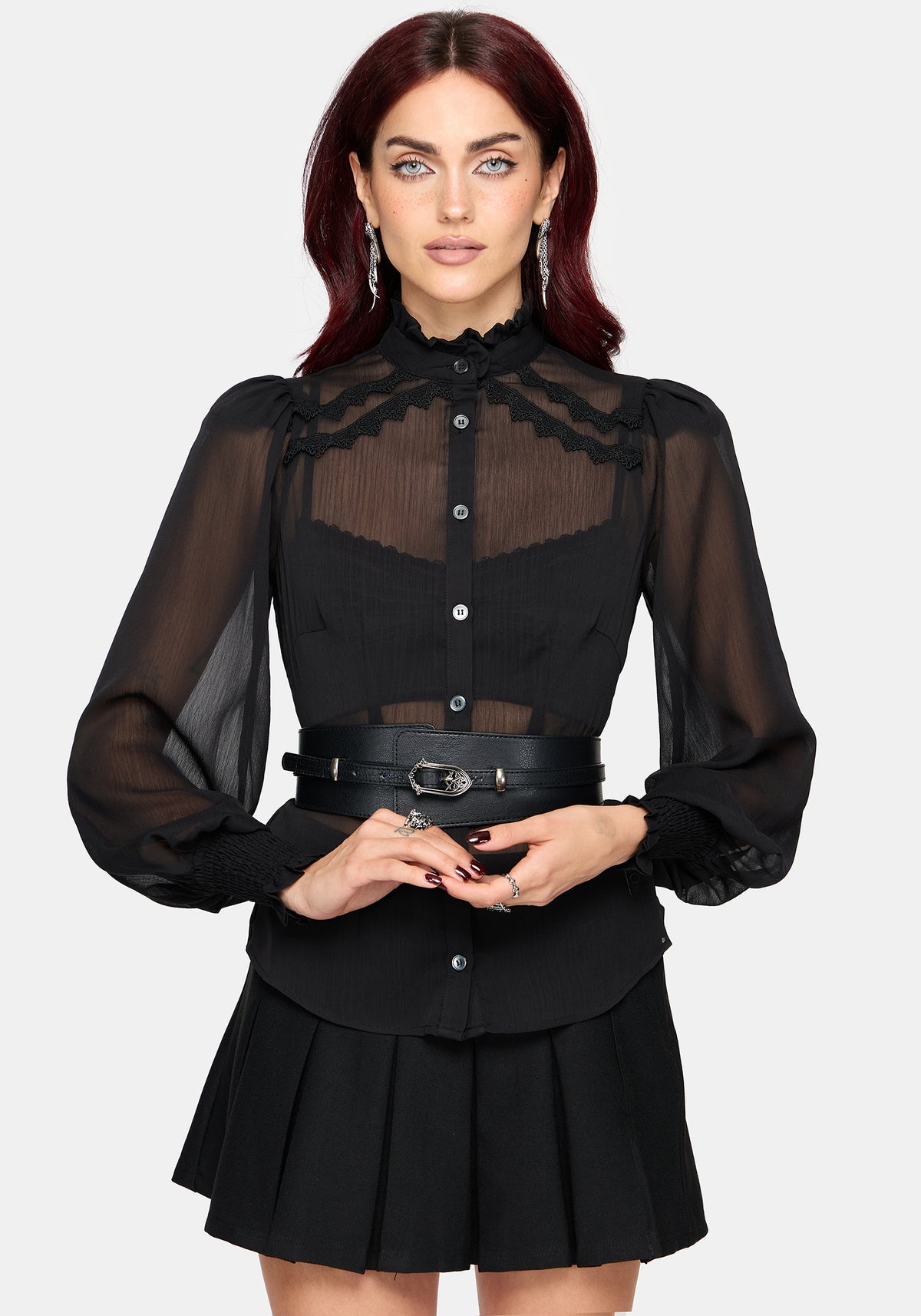 A woman modeling a black long-sleeve blouse with sheer mesh sleeves, a high ruffled collar, and a sheer yoke panel, paired with a matching pleated black skirt and a black belt with silver hardware, against a light gray background.