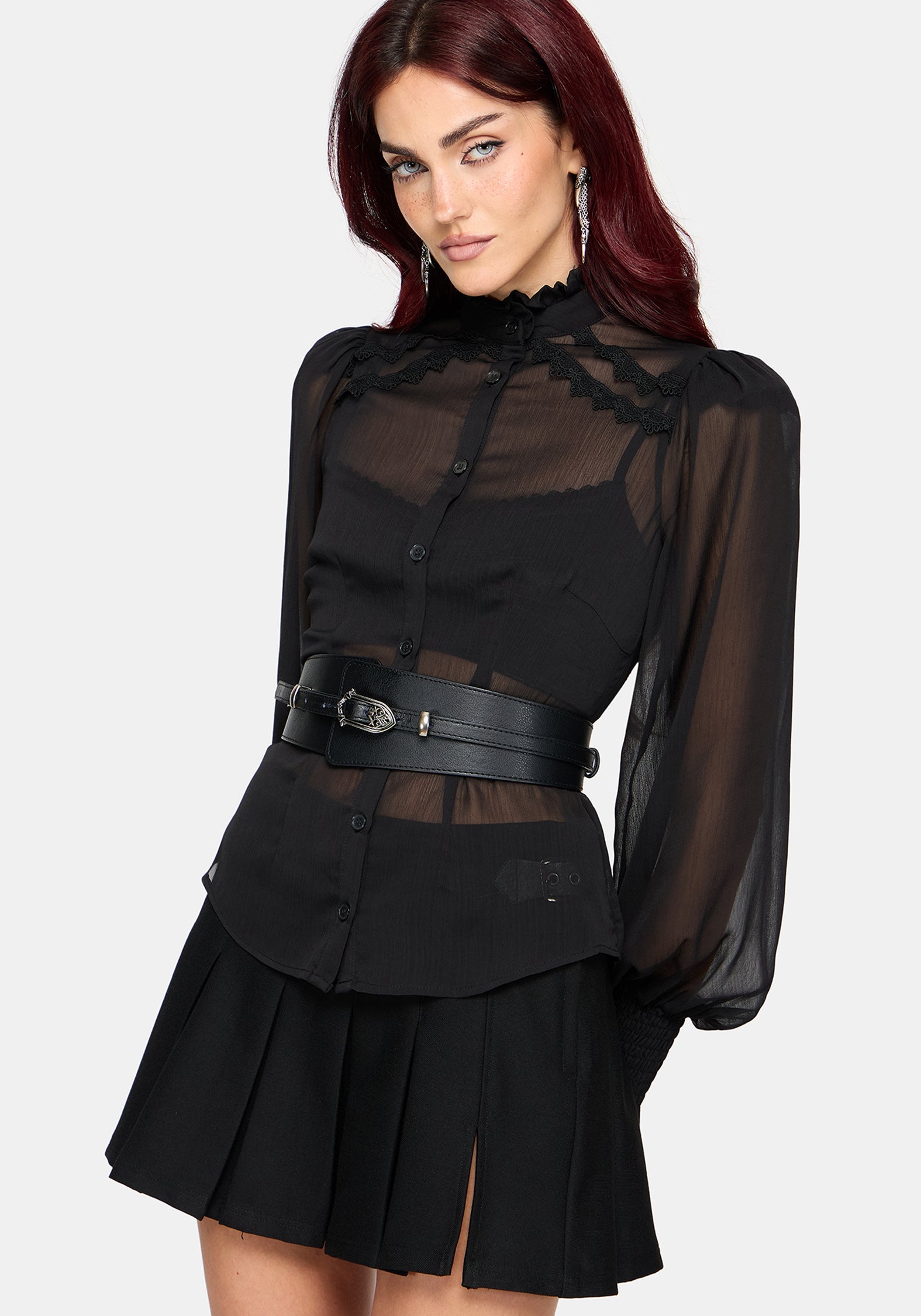 A model with dark red hair wearing a black sheer long-sleeve blouse with a lace-trimmed mock neck, a black pleated mini skirt, and a wide black belt with a silver buckle, set against a plain light gray background.