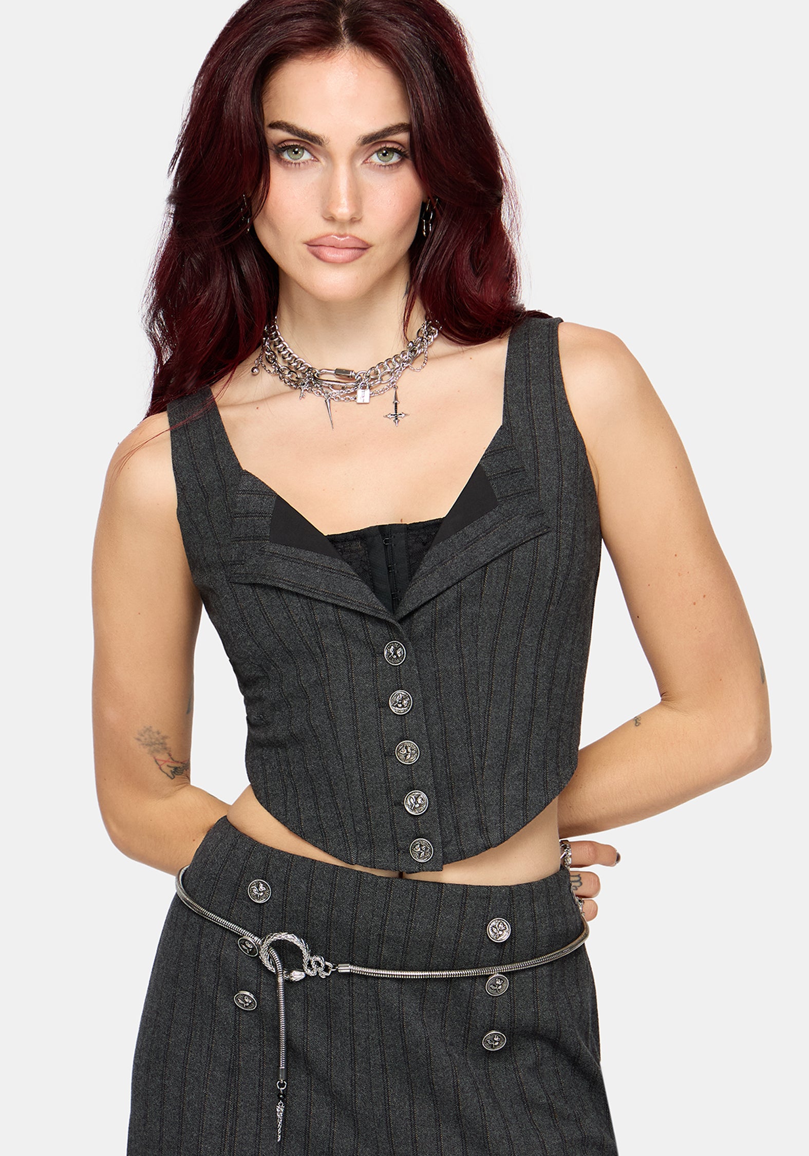 A woman modeling a dark gray pinstriped sleeveless vest corset with a button-front detail, paired with a matching skirt and silver accessories, against a light gray background.