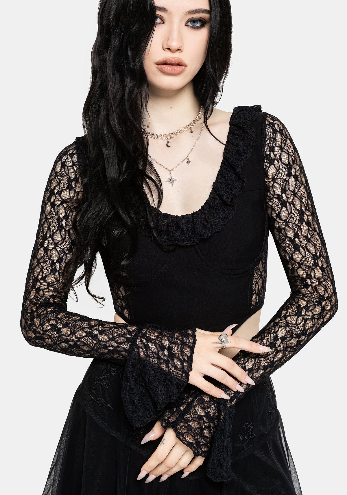 A woman modeling a black long-sleeve lace top with a ruffled scoop neckline and sheer lace sleeves, paired with a black skirt, against a light gray studio background.