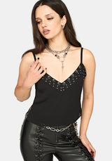 A woman modeling a black spaghetti-strap camisole top with silver stud embellishments along the V-neckline, paired with black lace-up pants and layered silver necklaces, against a light gray studio background.