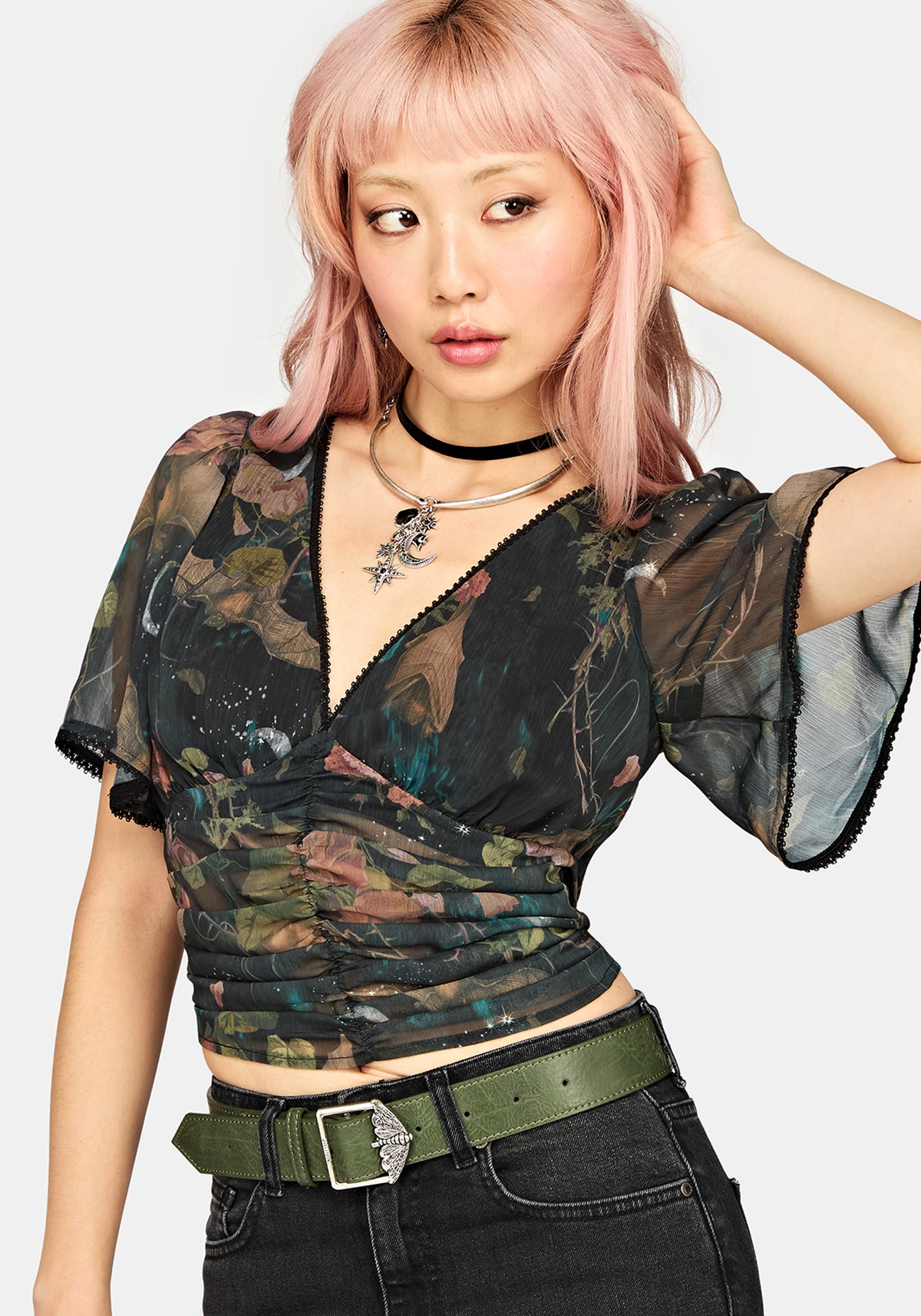 A model with pink hair wearing a short-sleeve wrap-style top with a dark camouflage pattern, paired with black jeans and an olive green belt, against a light gray background.