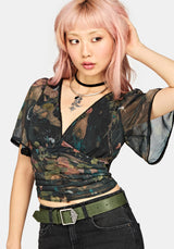 A model with pink hair wearing a short-sleeve wrap-style top with a dark camouflage pattern, paired with black jeans and an olive green belt, against a light gray background.