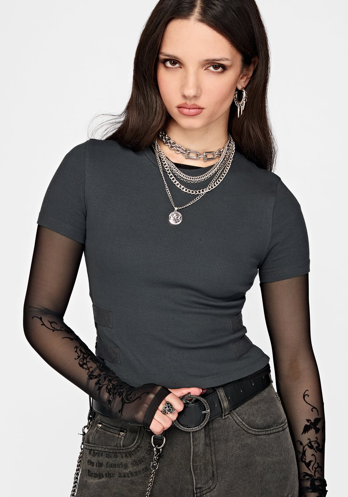 A woman modeling a dark charcoal gray short-sleeve ribbed t-shirt styled with layered silver necklaces, a black belt, and gray jeans against a light gray background.