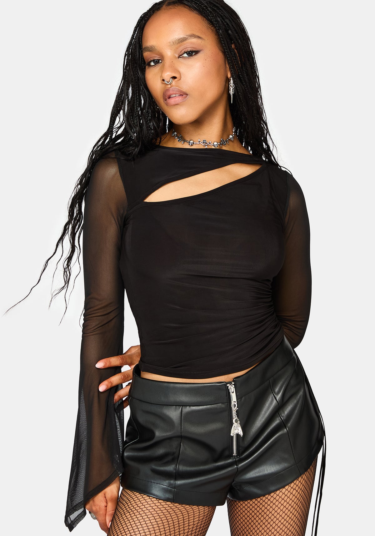 A woman modeling a black long-sleeve mesh top with a diagonal cutout at the chest and flared sleeves, paired with black faux-leather shorts, fishnet tights, and a silver choker necklace against a light gray background.