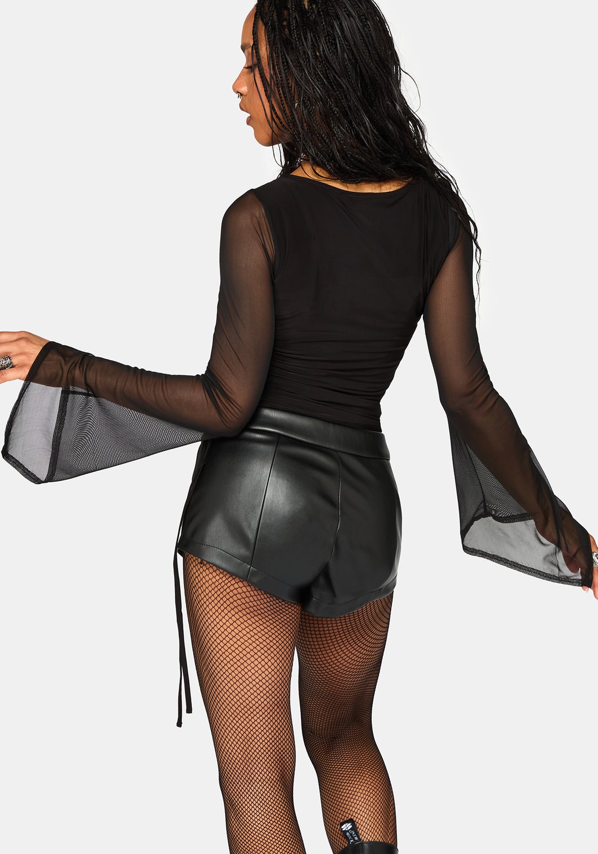 A woman shown from the back wearing a black sheer long-sleeve top with dramatic flared bell sleeves, black faux-leather shorts, and black fishnet tights against a light gray studio background.
