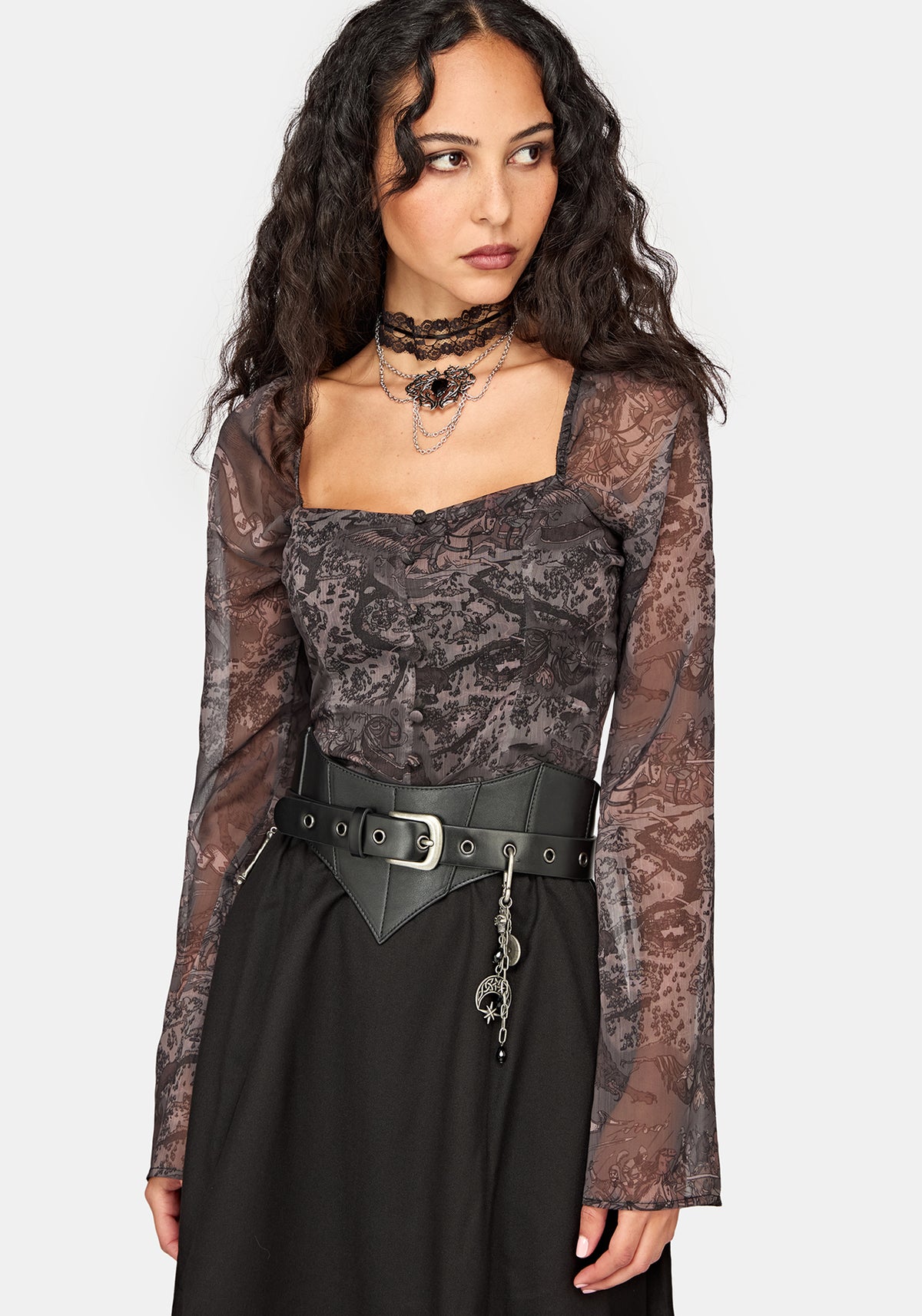 A model wearing a gothic-style outfit featuring a sheer black lace long-sleeve top with a dark floral pattern, a black leather belt with a silver buckle, and a black skirt, set against a plain light gray background.