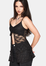 A woman modeling a black lace camisole top with thin straps, a ruched sweetheart neckline, and a side slit, paired with black pants against a light gray studio background.