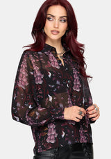 A woman with long, dark red hair models a sheer black long-sleeve blouse featuring a purple and pink floral pattern, paired with black lace-up pants, against a solid light gray background.