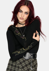 A model with dark red hair wearing a black long-sleeve top with a green and gold dragon graphic on the sleeve, paired with a plaid skirt and a silver belt.