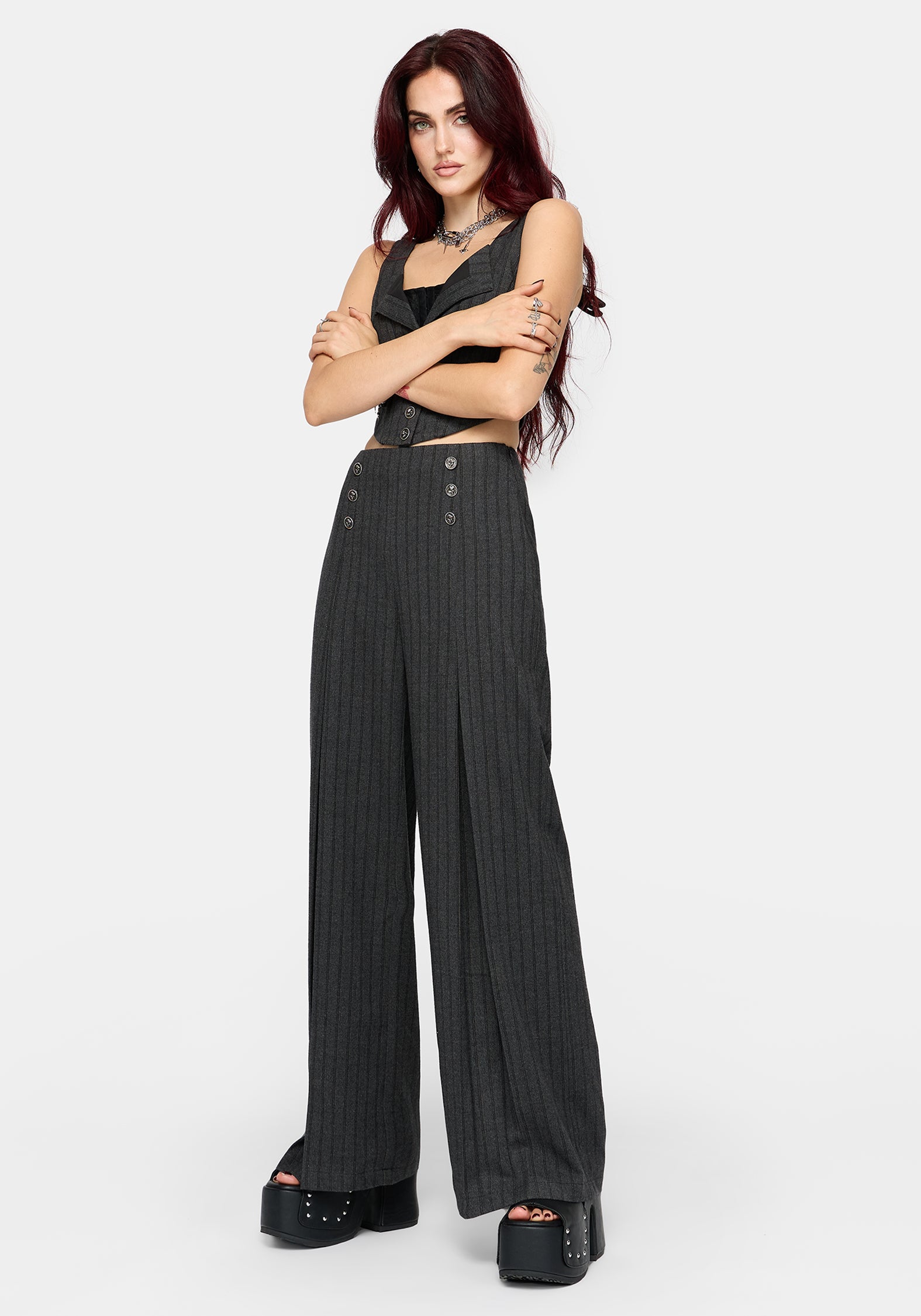 A model with dark red hair stands against a light gray studio background wearing a grey pinstriped crop top and matching high-waisted, wide-leg trousers with button details at the hips, paired with black platform sandals.