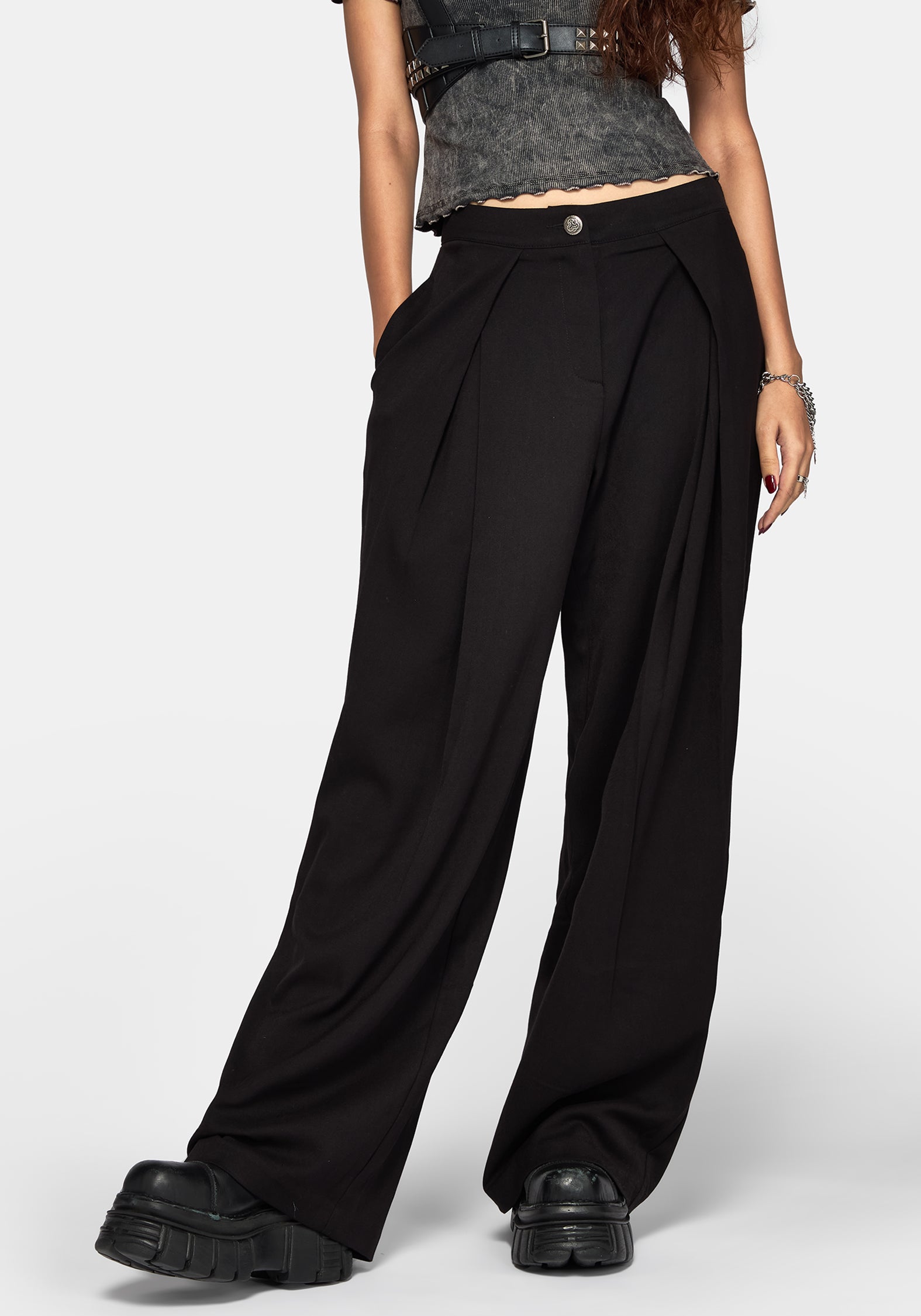 A model wearing black wide-leg pleated trousers with a button closure at the waist, styled with a gray textured crop top and chunky black platform shoes against a light gray studio background.