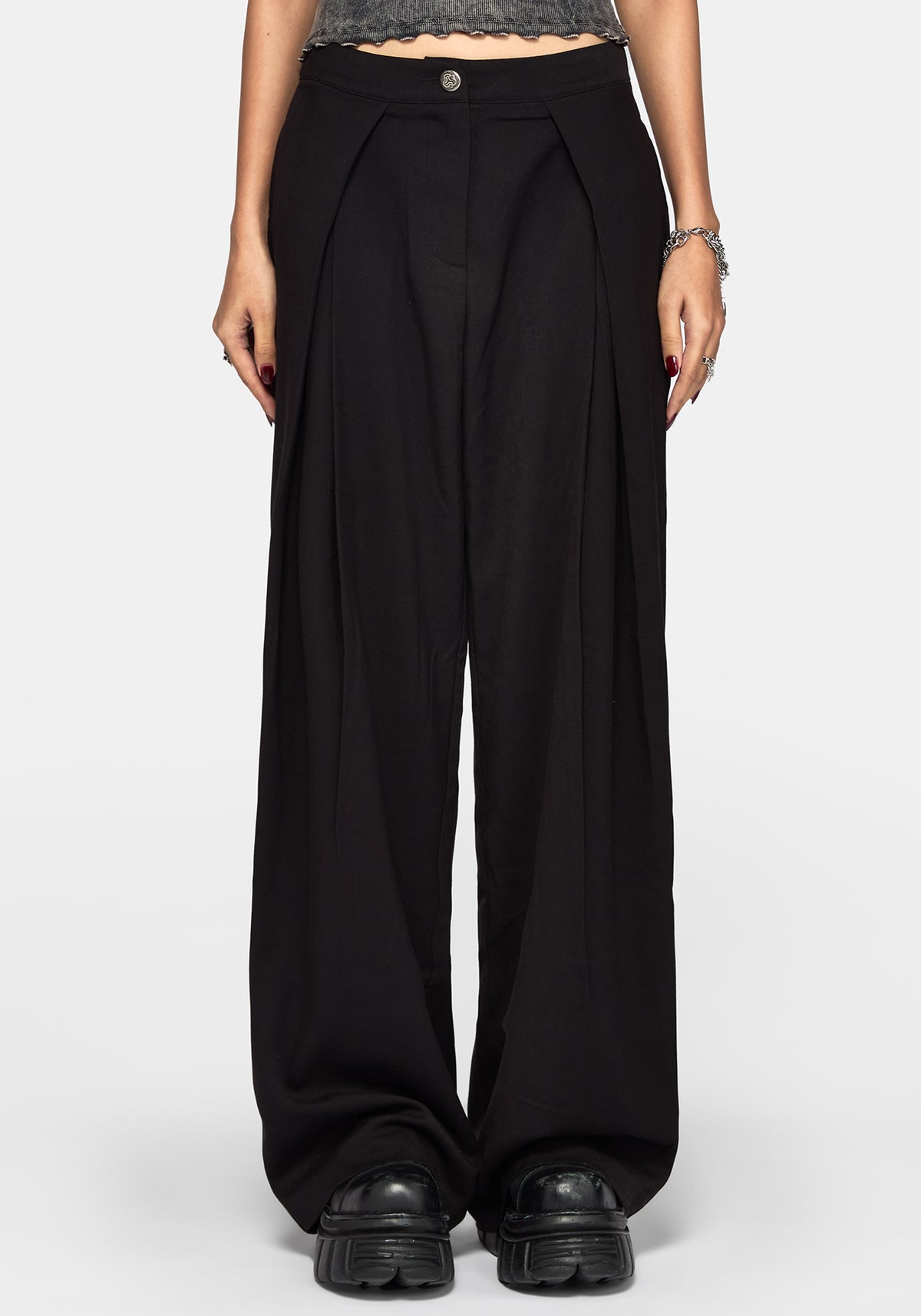 A model wearing black wide-leg pleated trousers with a button closure at the waist, styled with black platform shoes and a gray textured crop top against a light gray background.
