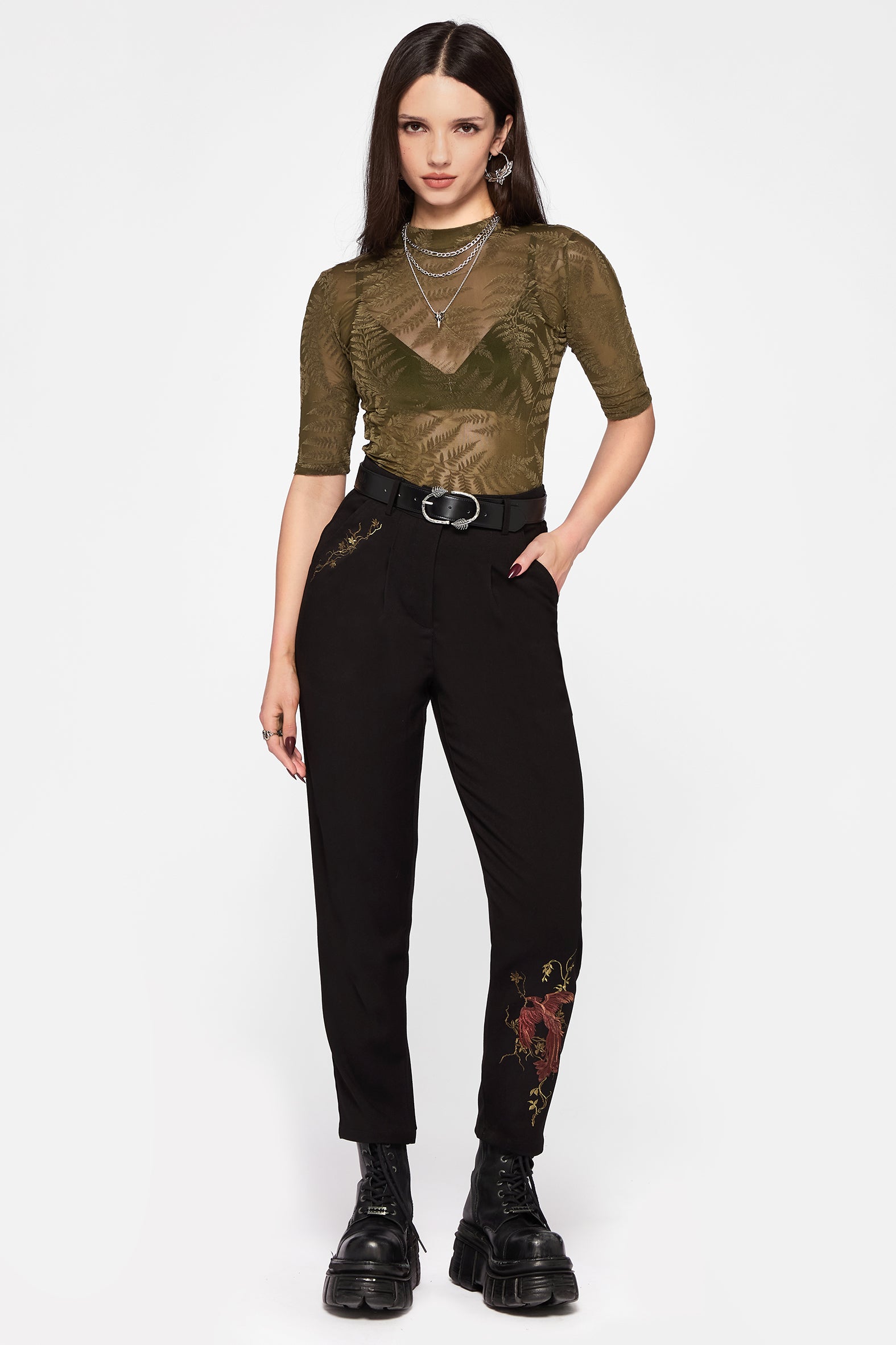 Woman wearing a green top and black pants with floral embroidery on a white background