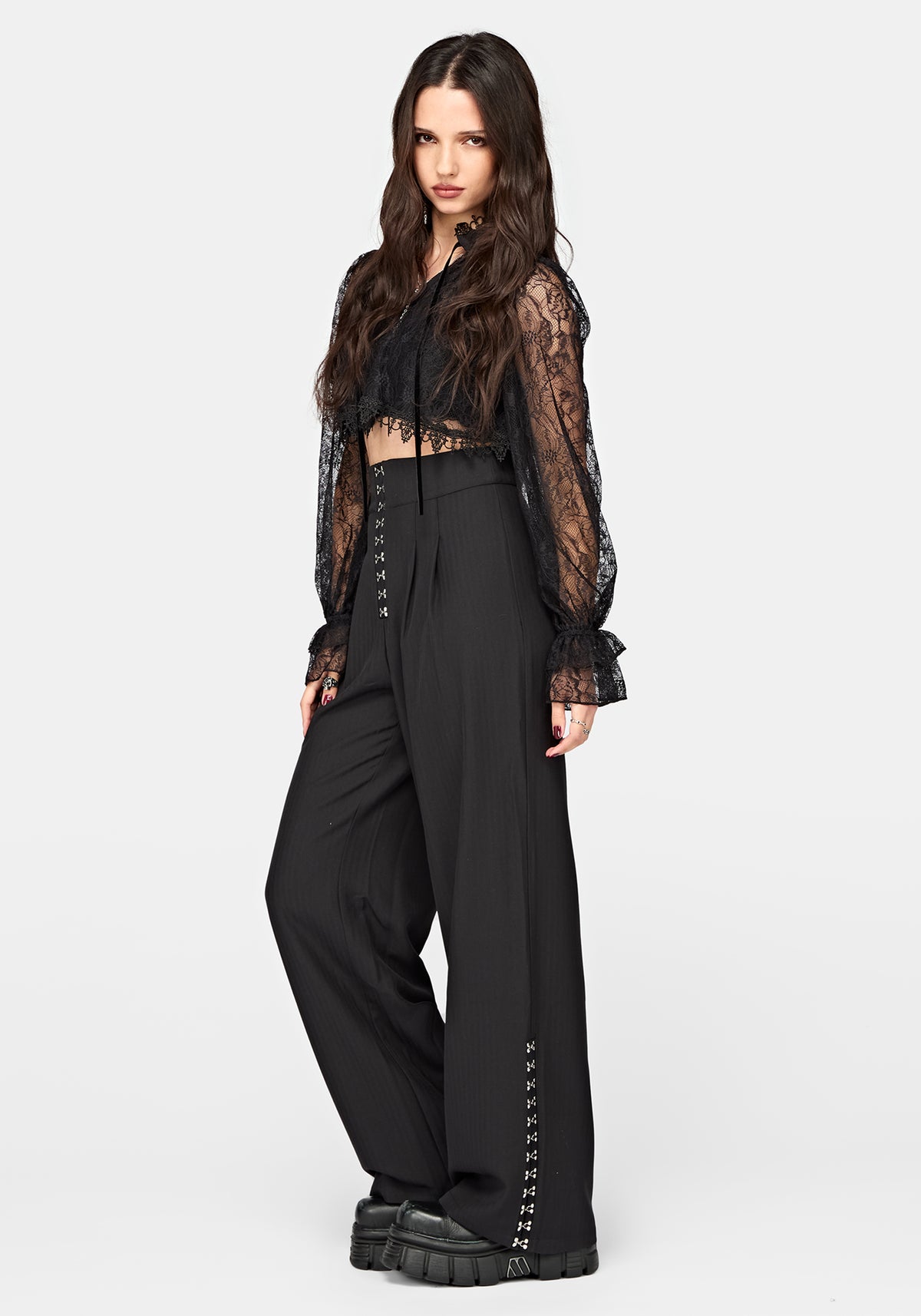 A model wearing a black gothic-style outfit consisting of a sheer black lace long-sleeve crop top, high-waisted black wide-leg trousers with lace-up front details, and chunky black platform shoes, set against a plain light gray studio background.
